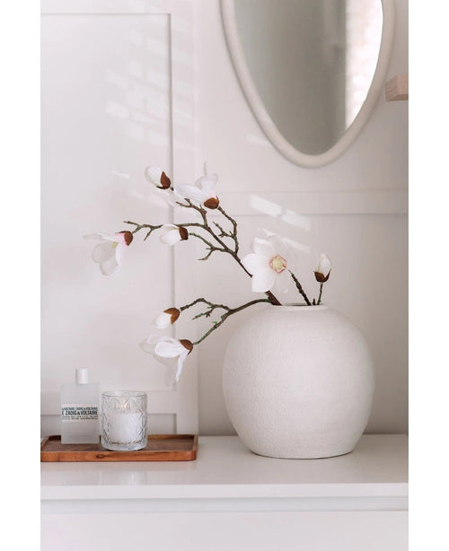 Une branche de magnolia artificielle Real Touch White 98 cm se dresse dans un vase rond blanc sur une surface blanche à côté d'une bougie en verre allumée et d'une petite bouteille sur un plateau en bois, dans une pièce lumineuse et minimaliste avec un miroir rond au-dessus.