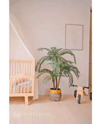 Une chambre d'enfant confortable avec un cadre de lit en bois clair, le palmier artificiel Cycas Classic 135 cm dans une jardinière jaune, un vélo jouet en bois avec le label MIA, des dessins au trait minimalistes sur le mur, du parquet en bois clair et une literie rose tendre pour plus de chaleur.