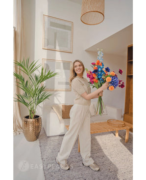 Une femme souriante vêtue de vêtements beige clair et de baskets se tient dans un salon lumineux et moderne avec des meubles en bois et des œuvres d'art encadrées, tenant un bouquet coloré. Le Palmier Kentia Artificiel 135 cm ajoute une touche tropicale à la pièce.