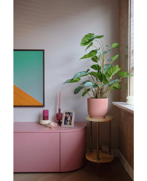 Un coin salon moderne avec un buffet rose, des bougies, une décoration murale géométrique, une photo encadrée et la plante artificielle Feuille de pommier de 90 cm - une grande plante à feuillage dans un pot rose sur un petit support en bois près de la fenêtre.