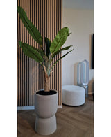 Le bananier artificiel de 150 cm, avec ses feuilles vertes luxuriantes, se dresse sur un plancher en bois près d'un mur nervuré avec des panneaux de bois. A proximité se trouve un pouf rond blanc et un ventilateur sans pales en arrière-plan.