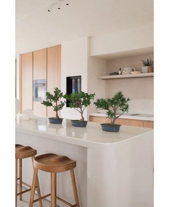 Une plante artificielle Bonsai Ficus de 40 cm se dresse sur un îlot de cuisine blanc avec deux tabourets en bois, entourés d'armoires en bois clair, de plans de travail blancs et d'étagères au décor minimaliste.