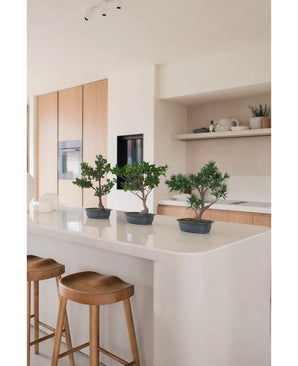 Une plante artificielle Bonsai Ficus de 40 cm se dresse sur un îlot de cuisine blanc avec deux tabourets en bois, entourés d'armoires en bois clair, de plans de travail blancs et d'étagères au décor minimaliste.