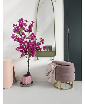 Un intérieur moderne avec le bougainvillier artificiel 125 cm violet dans un pot décoratif, un grand miroir avec un cadre vert, un repose-pieds rose avec base dorée et couverture rose, et une porte sombre contre un mur blanc éclatant.