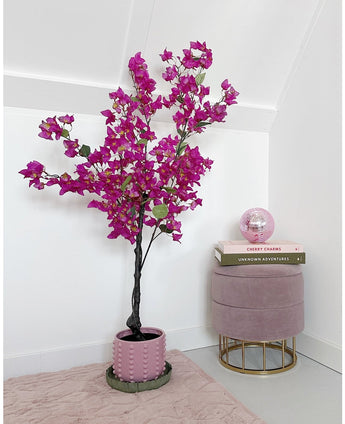 Le bougainvillier artificiel 125 cm violet avec des fleurs violettes éclatantes se trouve à côté d'un tabouret en velours rose avec des livres empilés sur le dessus et une boule de verre rose dans une pièce minimaliste et moderne aux murs blancs.