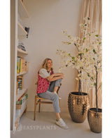 Une personne est assise souriante sur une chaise en bois à côté du bougainvillier artificiel 180 cm blanc dans un pot doré avec texture. À gauche se trouve une étagère avec des livres et de la décoration ; la lumière du jour entre par une fenêtre sur la droite.