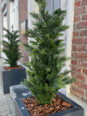 Un gros plan de la plante artificielle Conifer UV 120 cm, une plante conifère artificielle verte dans une jardinière rectangulaire noire avec un paillis d'écorce brune, exposée sur un trottoir près d'un bâtiment en brique ; un autre planteur de ce type est en arrière-plan.