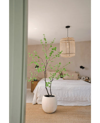 Une chambre minimaliste avec la plante Artificielle Enkianthus 180 cm en grande plante en pot, un lit avec une literie blanche, une suspension tressée, des murs beiges et un tapis en fibres naturelles pour une ambiance simple et aérée.