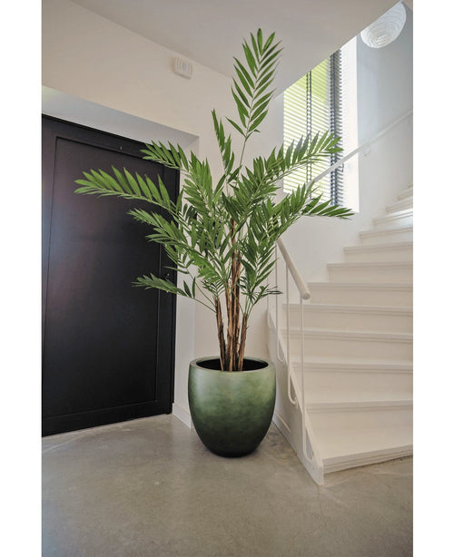 La plante artificielle Hawaii Parlour 180 cm, un grand palmier artificiel d'intérieur aux feuilles vertes luxuriantes, se dresse sur un sol gris dans un hall moderne, près d'un escalier blanc et d'une porte noire.