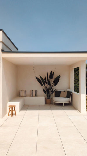 Une terrasse moderne et minimaliste avec du carrelage couleur crème, un canapé intégré avec coussins rayés, une plante artificielle Heliconia 210 cm pour une touche luxueuse, un petit tabouret en bois et une chaise ronde avec coussins sous un toit plat sur un ciel bleu clair.