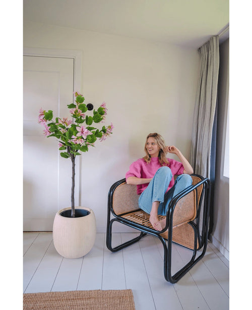 Une femme vêtue d'un pull rose et d'un jean bleu est assise pieds nus sur une chaise en rotin à côté de la plante artificielle Judas 150 cm, un arbre artificiel à fleurs roses, dans une pièce lumineuse et moderne aux murs et au sol blancs.
