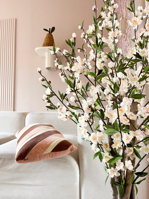Un salon cosy avec un canapé blanc, un coussin rayé rose et marron et la branche fleurie de cerisier artificiel de 120 cm crème au premier plan. Une plante en pot repose sur une étagère murale contre un mur rose pâle.