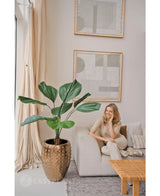 Une femme se détend sur un canapé lumineux dans un salon moderne aux couleurs neutres. À côté d'elle se trouve la plante artificielle Lucuala Grandis de 120 cm dans un pot, tandis que des œuvres d'art abstraites sont suspendues au-dessus et que la lumière naturelle illumine l'espace.