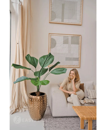 Une femme se détend sur un canapé lumineux dans un salon moderne aux couleurs neutres. À côté d'elle se trouve la plante artificielle Lucuala Grandis de 120 cm dans un pot, tandis que des œuvres d'art abstraites sont suspendues au-dessus et que la lumière naturelle illumine l'espace.