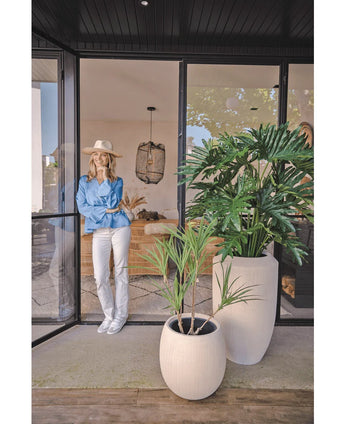 Une femme portant un chapeau, un chemisier bleu et un pantalon blanc s'appuie contre une porte vitrée. Au premier plan se trouve la plante artificielle Philodendron 125 cm. À l’intérieur, vous pouvez voir un espace de vie confortable et moderne.
