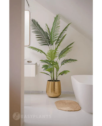 La plante artificielle Phoenix 180 cm dans un pot doré se trouve à côté d'une baignoire et d'un lavabo blancs dans une salle de bain moderne et minimaliste aux murs et au sol blancs. A proximité se trouve un tapis rond tissé sur le sol.