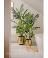 Une plante artificielle Phoenix de 180 cm avec des feuilles vertes luxuriantes dans un pot doré brillant se trouve à côté d'une baignoire blanche et d'un tapis tissé beige dans une salle de bain moderne et minimaliste, complétée par un miroir et une fenêtre pour un look serein.
