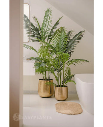 Une plante artificielle Phoenix de 180 cm avec des feuilles vertes luxuriantes dans un pot doré brillant se trouve à côté d'une baignoire blanche et d'un tapis tissé beige dans une salle de bain moderne et minimaliste, complétée par un miroir et une fenêtre pour un look serein.