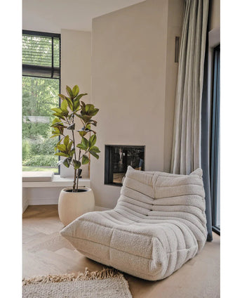 Une chaise longue confortable et texturée de couleur crème se trouve à côté d’une cheminée moderne et d’une grande fenêtre. A côté se trouve l'arbre Yulania artificiel de 150 cm dans un pot rond blanc. Les parquets en bois clair et la douce lumière du soleil créent une atmosphère calme et accueillante.