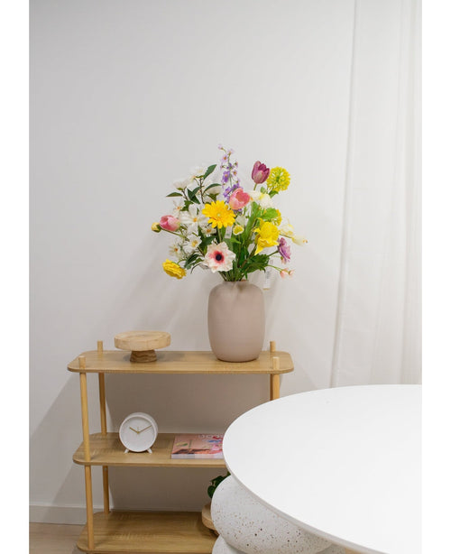 Un vase beige avec des fleurs artificielles de renoncule Real Touch jaune de 60 cm se trouve sur une étagère en bois clair à côté d'une table ronde blanche. Sur l'étagère se trouvent également une petite horloge blanche, un plateau en bois et un livre contre un mur blanc uni.