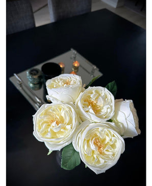 Le Bouquet de Roses Artificielles "Carmen" Real Touch Blanc Crème 36cm, avec cinq roses de couleur crème, repose sur une table sombre. Derrière elle se trouve une plate-forme rectangulaire en miroir avec des bougies et un décor, avec des chaises grises partiellement visibles.