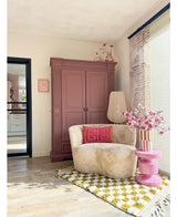 Un coin lecture confortable comprend une chaise crème, des coussins roses, un tapis à motifs jaunes et une armoire mauve. La Branche Artificielle Magnolia à petites fleurs 89 cm rose se dresse sur une table d'appoint rose, tandis que de grandes fenêtres remplissent cet espace invitant d'une lumière douce.