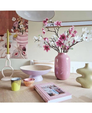 Une pièce moderne comprend une table dressée avec une branche artificielle de magnolia à petites fleurs de 89 cm rose dans un vase rose, des bougeoirs de couleur pastel, des bols, des tasses, un vase sculptural vert, un livre rose et des œuvres d'art florales au mur.