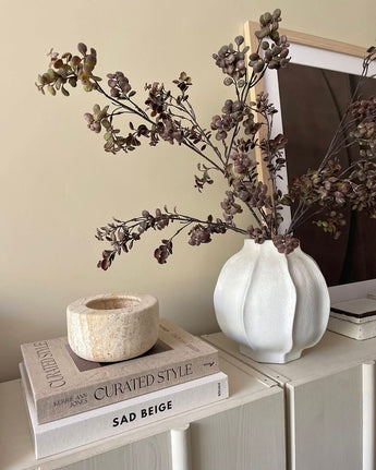 Sur une armoire lumineuse se trouve un vase blanc rempli de branche artificielle d'eucalyptus de 110 cm marron, à côté d'une pile de livres et d'un bougeoir en pierre, avec une photo encadrée en arrière-plan.