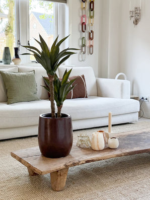 Un salon moderne avec un canapé blanc, des coussins verts et marron, une table basse en bois décorée de bougies et d'agave artificielle 76 cm, et une décoration murale en maille chaîne. La lumière naturelle pénètre par les fenêtres derrière le canapé.