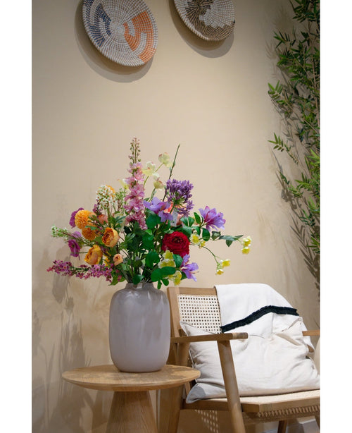 Un vase blanc avec cinq branches artificielles de Ranunculus orange vif x5 fleurs Real Touch 62 cm se trouve sur une table en bois à côté d'une chaise en bois avec un coussin blanc et une couverture ; des paniers tressés sont suspendus à un mur beige et des branches vertes apparaissent à proximité.