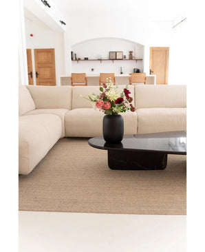Un salon moderne avec un canapé beige, un tapis neutre et une table basse noire avec le Vase Artic Black Ø21 x H29 cm à fleurs roses, blanches et rouges. Des étagères et des portes en bois apparaissent en arrière-plan.