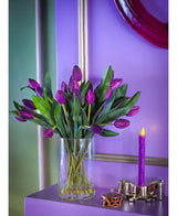 Le bouquet artificiel en soie Tulipes dans un vase violet présente des tulipes violettes vibrantes avec des feuilles vertes dans un vase en verre transparent, exposées sur une surface violette à côté d'une bougie allumée et d'un récipient décoratif, sur un fond bleu lavande avec des accents métalliques.
