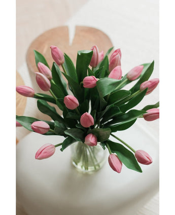 Le bouquet de tulipes en soie artificielle dans un vase rose montre des tulipes roses artificielles avec des feuilles vertes, disposées dans un vase en verre, affichées sur une surface blanche avec un fond clair légèrement décoloré, vues d'en haut.