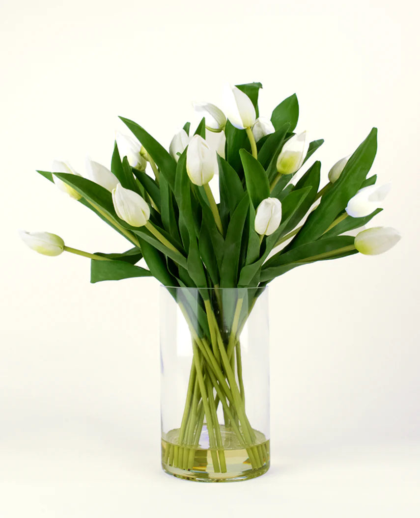 Le bouquet artificiel en soie Tulipes dans un vase Blanc présente des tulipes blanches avec des feuilles et des tiges vertes dans un vase en verre transparent, sur un fond clair simple.