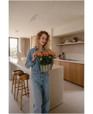 Une femme aux cheveux blonds bouclés, vêtue d'une chemise en jean et d'un jean, se tient dans une cuisine moderne, tenant une plante UV artificielle marguerite de 31 cm avec des fleurs orange vif. À côté d’elle se trouvent deux tabourets en bois près de l’îlot de cuisine.