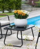 Une table de patio noire avec une plante de marguerite artificielle UV de 31 cm – un pot blanc rond avec des marguerites orange et blanches éclatantes – se dresse à côté d'une piscine, entourée de chaises de jardin et avec de la verdure en arrière-plan.