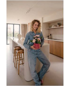 Une femme en jeans se tient dans une cuisine moderne, souriante et tenant dans ses mains la plante artificielle Gerbera UV rose 28 cm – un petit pot de fleur rose avec des fleurs roses et blanches. Au fond, on aperçoit deux tabourets en bois et une grande fenêtre.