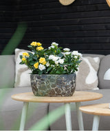 Une Plante Artificielle Patio Rose 35 cm crème UV se dresse sur une table en bois devant un canapé gris avec des coussins à motifs, sur fond de murs en bois sombre.