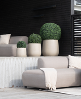Quatre Buis Artificiels de style romain UV D40cm, ronds et beiges, avec des arbustes verts bien entretenus, sont placés sur une terrasse moderne avec des chaises gris clair, une couverture beige et un fond en bois foncé pour un look minimaliste.
