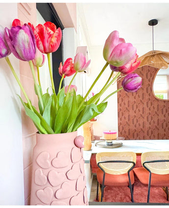 Un vase rose avec des cœurs en relief contient la fleur artificielle French Tulip 60 cm rouge sur une table à manger. Derrière, se trouvent des chaises avec dossier en osier, un gâteau sur la table et un miroir sur un mur marron à motifs sous une suspension tressée.