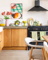 Une cuisine lumineuse avec des armoires en bois, une cuisinière noire, des serviettes vertes, de la vaisselle colorée et une photo de pâtisserie encadrée sur une table à carreaux prend un nouveau look grâce à la Fleur Artificielle Perruche Française Tulipe 76 cm rouge/jaune. Une chaise blanche confortable se trouve à côté d’une table à manger en bois clair.