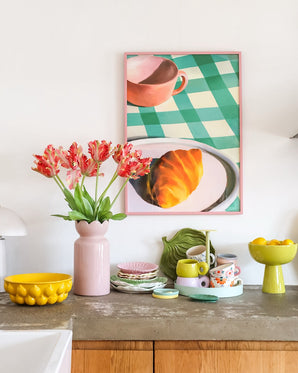 Un plan de travail avec toutes sortes de vaisselle colorée et une fleur artificielle Tulipe Perruche Française de 76 cm en rouge/jaune dans un vase rose. Au-dessus est accrochée une photo encadrée d’un croissant et d’une tasse de thé sur une nappe à carreaux verts.