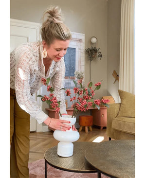 Une femme aux cheveux blonds en chignon dispose le Kunsttak Berries 64 cm rose dans un vase blanc sur une table ronde dans un salon cosy aux tons terre et au mobilier moderne.