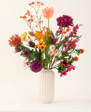 Un vase en céramique blanche et nervurée présente le bouquet de fleurs artificielles en soie Magnus di Lambo - un arrangement de fleurs artificielles orange vif, roses, violettes et jaunes avec des feuilles vertes sur un fond clair.