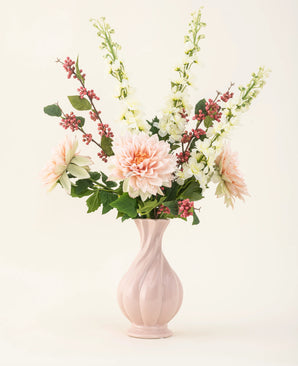 Un vase en céramique rose contient le bouquet de fleurs artificielles en soie Morning Whisper, avec des dahlias rose pâle, des mufliers blancs, des feuilles vertes et des grappes de petites baies rouges, sur un fond uni blanc cassé.