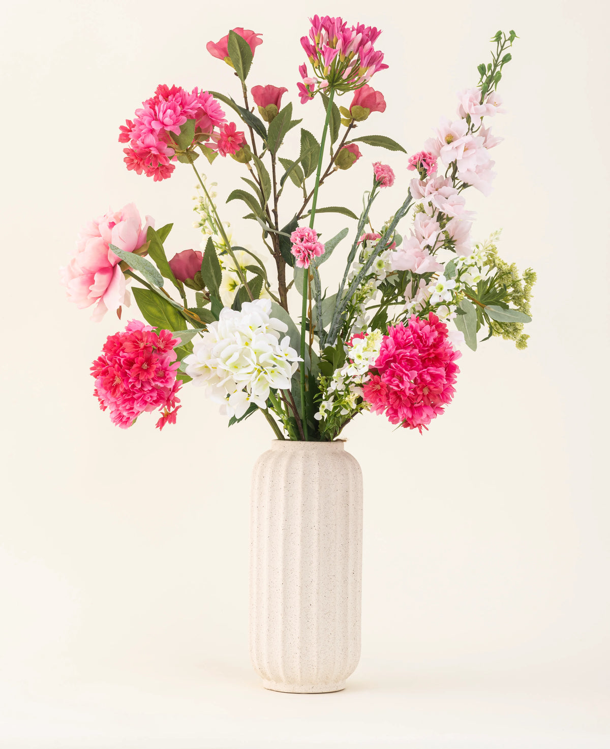 Un vase blanc texturé montre le bouquet de fleurs artificielles en soie Blush Breeze – des fleurs en soie roses, blanches et violet clair avec des feuilles vertes – sur un fond clair uni.