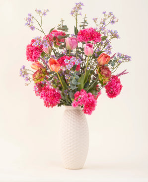 Un vase à structure blanche et le bouquet de fleurs artificielles en soie Soft Spring avec des fleurs rose vif, des tulipes orange et des fleurs bleu violet apportent le printemps dans votre maison sur un fond uni clair.