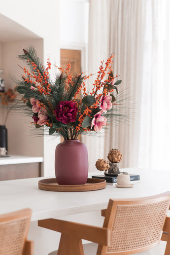 Le bouquet de fleurs artificielles en soie Cosy Winter dans un vase mauve, avec des fleurs roses, des baies oranges et des branches de pin, orne un plateau rond en bois sur une table blanche pour un look automne/hiver cosy et moderne avec des chaises en bois et des rideaux beiges.