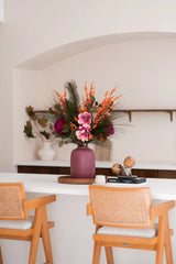 Un vase mauve avec le bouquet de fleurs artificielles en soie Cosy Winter - parfait pour la décoration automne-hiver - se dresse sur un comptoir blanc entre des chaises en bois, avec des livres, des artichauts, des étagères et un pot blanc avec des branches à proximité.