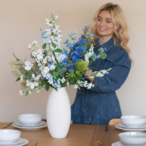 Une femme souriante vêtue d'une robe en jean bleue dispose le bouquet de fleurs artificielles en soie Cool Breeze dans un vase blanc structuré sur une table à manger garnie de vaisselle blanche et de couverts dorés.